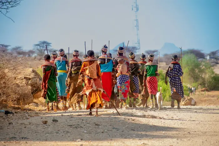 A pastoralist community walking a dirt road past a mobile network mast